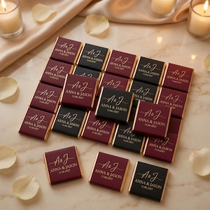 May include: A collection of square chocolate bars wrapped in burgundy and black paper. Each bar features gold foil text reading A & J, ANNA & JASON, and the date 11.06.2027. The bars are arranged in a grid pattern on a light surface with scattered petals.