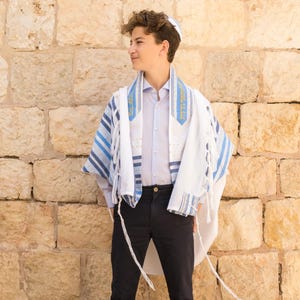 May include: A white tallit prayer shawl featuring blue and silver striped patterns along the edges, worn over a light blue button-down shirt and dark trousers. The shawl includes Hebrew text embroidered in gold on the atarah neckband.
