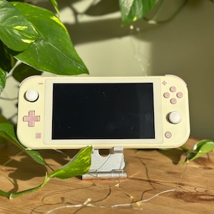 Op de afbeelding: Een draagbare videogameconsole in een lichtgele afwerking met een zwart scherm. Het apparaat is voorzien van een richtingspad en actieknoppen in een gedempte roze tint, samen met twee witte analoge sticks, gemonteerd op een zilveren metalen standaard.