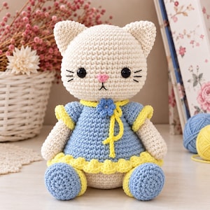 Puede incluir: Una muñeca de gato tejida a ganchillo de color crema con ojos de seguridad negros, nariz rosa y bigotes bordados. El gato lleva un vestido de ganchillo azul con ribete amarillo, un cordón amarillo y una pequeña flor azul en el cuello.