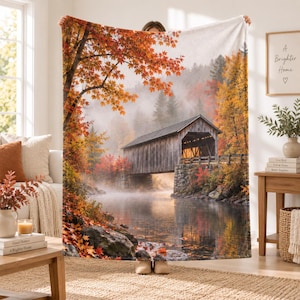 May include: A rectangular fleece blanket featuring a printed landscape of a wooden covered bridge over a calm river, surrounded by autumn trees with vibrant orange and red foliage, set against a misty forest background with rocky banks.