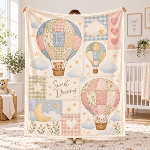 May include: A cream-colored throw blanket featuring a patchwork design of hot air balloons, clouds, stars, hearts, and a crescent moon. The blanket displays a teddy bear and a rabbit in baskets, with the text Sweet Dreams printed in the center.