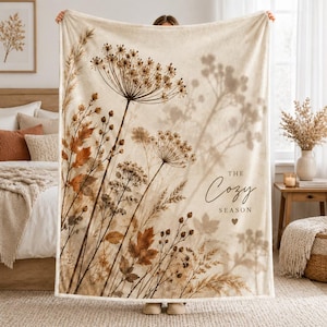 Può includere: Una coperta color crema con una stampa botanica di fiori selvatici essiccati, erbe e foglie autunnali in tonalità marroni e beige. Il testo "THE Cozy SEASON" è stampato in un carattere corsivo marrone scuro con un piccolo cuore.