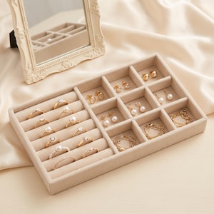 May include: A beige velvet jewellery organiser tray featuring multiple compartments for rings, earrings, and bracelets. The tray contains various gold-toned rings with pearls and clear stones, gold stud earrings, and delicate gold chain bracelets.