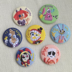 Può includere: Sette bottoni rotondi ricamati con personaggi dei cartoni animati. I bottoni mostrano i personaggi etichettati come Ragatha, Zooble, Kinger, Caine, Jax, Pomni e Gangle, ognuno con colori distinti e nomi ricamati su vari sfondi in tessuto.