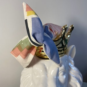 Può includere: Un fiocco di stoffa colorato legato attorno a una scultura di testa di leone in ceramica bianca. Il fiocco è fatto di un tessuto a fantasia con forme geometriche e strisce in tonalità di blu, rosa, verde, giallo e rosso.