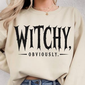 May include: A beige crewneck sweatshirt featuring the black text WITCHY, OBVIOUSLY. printed in a stylized, jagged font across the chest. The garment has long sleeves and a ribbed neckline, providing a simple and clean look for casual wear.