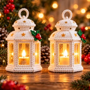 May include: Two white crocheted lanterns with hexagonal frames, each featuring a top ring handle, small gold star accents, and a green leaf and red berry embellishment. Inside each lantern sits a lit white pillar candle.