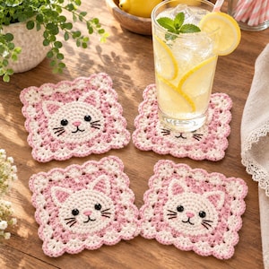 May include: A set of four square crochet coasters featuring a white cat face with black eyes, a pink nose, and whiskers, surrounded by a scalloped pink and white border. The coasters are crafted with a textured yarn finish and a consistent design.