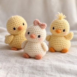 May include: Three small crocheted plush toys shaped like chicks. Two are bright yellow with orange beaks and feet, while the center one is cream-colored with a small pink bow on its head, an orange beak, and orange feet. All have black eyes.