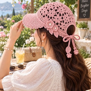 May include: A light pink crochet baseball cap featuring an intricate floral pattern throughout the crown and brim. The cap is adorned with two layered crochet flowers with pearl centers on the side and a bow closure at the back with dangling leaf and flower charms.