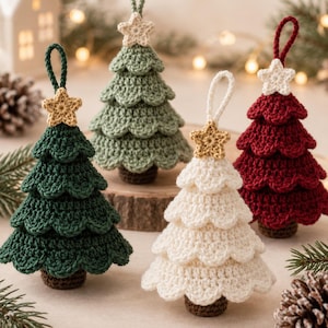 Op de afbeelding: Vier gehaakte kerstboomornamenten in donkergroen, saliegroen, crème en dieprood. Elke boom heeft drie geschulpte lagen, een bruine stam en een kleine gehaakte ster aan de top met een bijpassend garenlusje om aan een tak op te hangen.