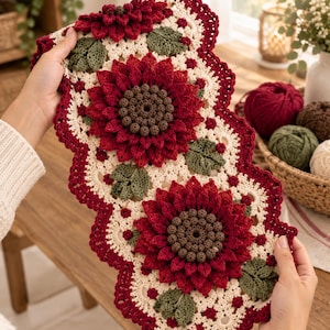 Op de afbeelding: Een rechthoekige gehaakte tafelloper met drie grote, driedimensionale donkerrode zonnebloemen met bruine harten. De bloemen zijn omringd door crèmekleurig kantachtig haakwerk en kleine groene blaadjes. De randen zijn donkerrood en geschulpt.