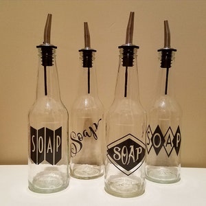 May include: Four clear glass bottles with black metal spouts and labels that say "SOAP" in different styles. The bottles are on a white surface.