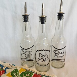 May include: Three clear glass bottles with black and white labels. The labels read "French Kitchen", "Dish Soap", and "Dish Soap". The bottles have silver tops with a spout.