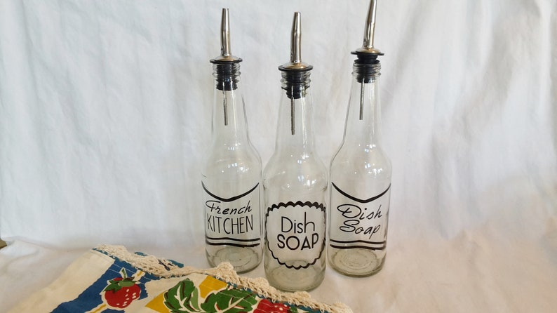 May include: Three clear glass bottles with black and white labels. The labels read "French Kitchen", "Dish Soap", and "Dish Soap". The bottles have silver tops with a spout.