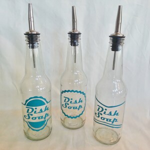 May include: Three clear glass bottles with silver spouts and black tops. Each bottle has a teal blue label that says "Dish Soap" in a vintage font.