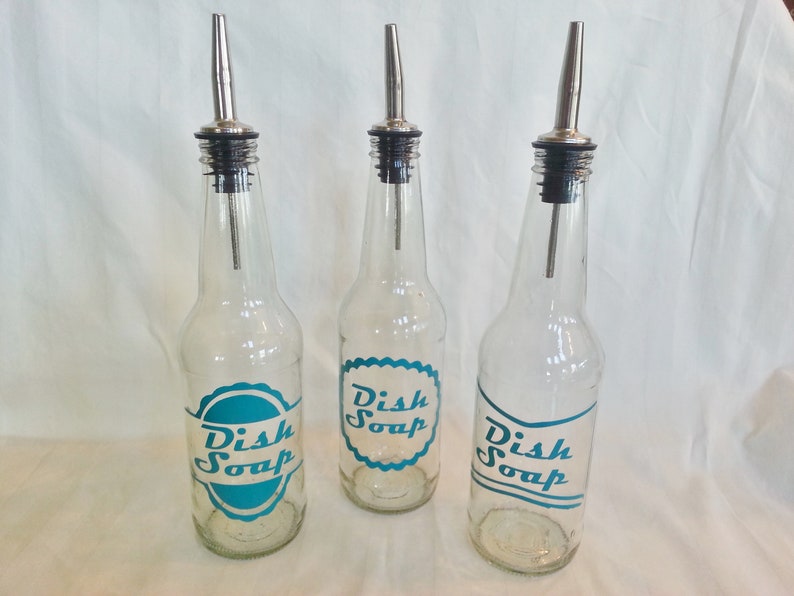 May include: Three clear glass bottles with silver spouts and black tops. Each bottle has a teal blue label that says "Dish Soap" in a vintage font.