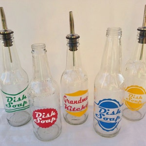 May include: Five clear glass bottles with metal pour spouts and labels. The labels are in different colors and say "Dish Soap", "Grandma's Kitchen", and "Dish Soap".