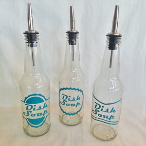 May include: Three clear glass bottles with metal spouts and black stoppers. Each bottle has a teal label with the words "Dish Soap" in white script. The bottles are arranged in a row against a white background.