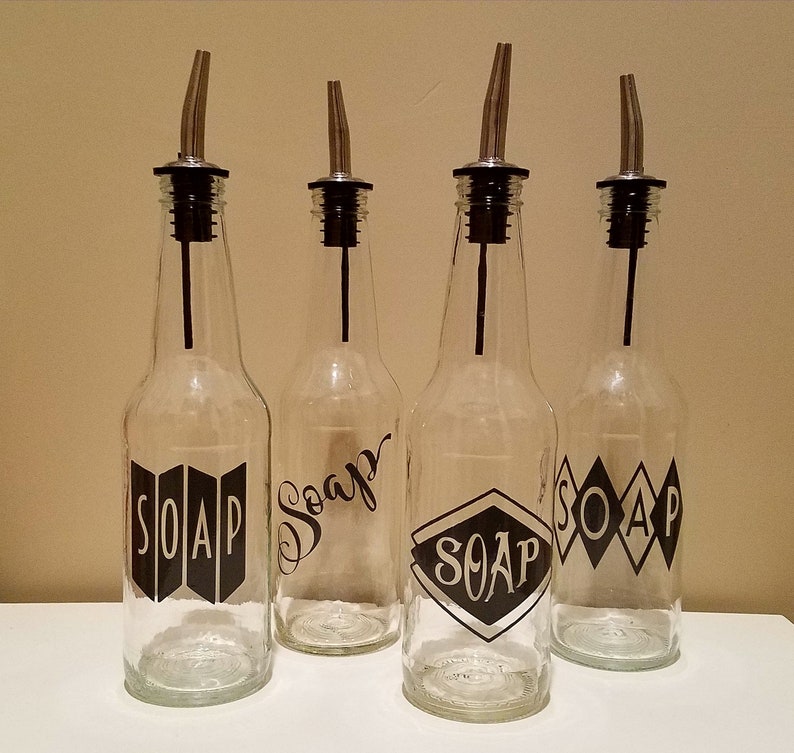 May include: Four clear glass bottles with black metal spouts and labels that say "SOAP" in different styles. The bottles are on a white surface.