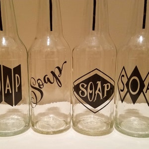 May include: Four clear glass bottles with black labels that say "SOAP" in different styles. The bottles are all the same size and shape.