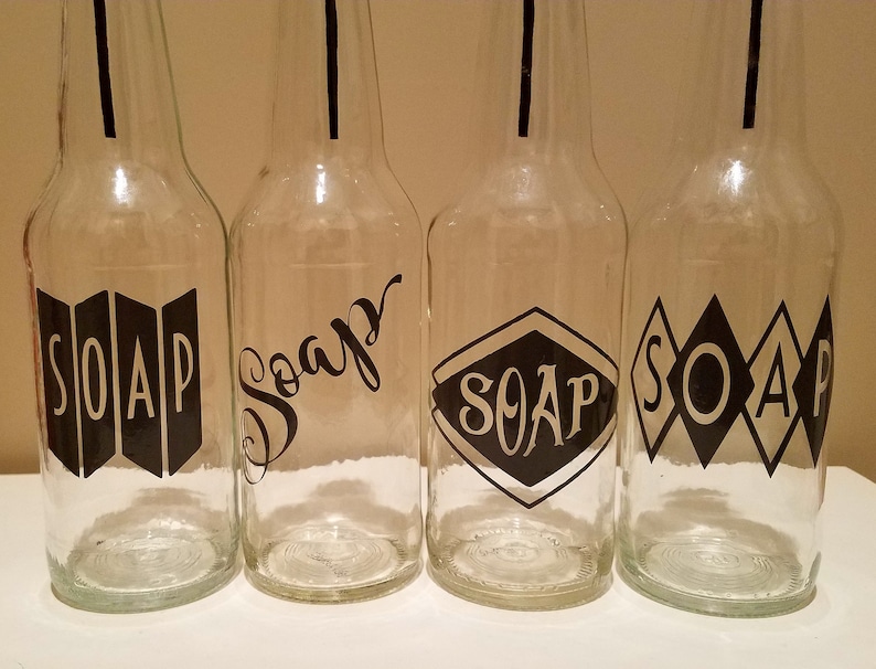 May include: Four clear glass bottles with black labels that say "SOAP" in different styles. The bottles are all the same size and shape.