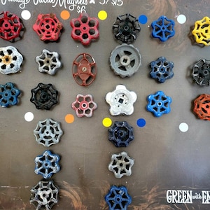 May include: A collection of 22 vintage valve handle magnets in various colors, including red, blue, green, yellow, and silver. The magnets are arranged in a grid pattern on a brown surface. The text "GREEN with ENVY" is visible in the bottom right corner of the image.