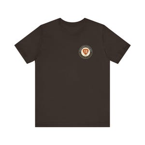 以下が含まれることがあります： 左胸に円形のロゴが入ったダークブラウンの半袖Tシャツ。ロゴには数字の18が描かれたオレンジ色の盾があり、その周囲を円形の枠で囲まれたEIGHTEEN & ROLLINGおよびBUILT FOR THE LONG HAULというテキストが囲んでいます。