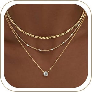 May include: A layered gold-tone necklace set consisting of three distinct chains. The top chain is a classic curb link, the middle chain features small metallic accents, and the bottom chain is a delicate rope style with a single round clear stone.