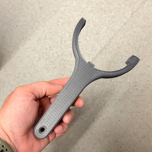 Peut inclure: Un outil en plastique gris imprimé en 3D doté d'un long manche plat avec un trou circulaire à la base et d'une tête large, incurvée et en forme de Y avec deux dents saillantes au sommet, présentant une texture en couches visible sur l'objet.