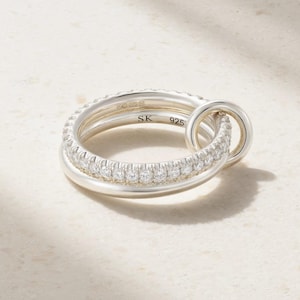 Op de afbeelding: Een zilveren ring bestaande uit een gladde band verbonden met een tweede band bezet met een rij heldere, rondgeslepen stenen. De binnenzijde van de ring is gegraveerd met de letters SK en het getal 925, wat duidt op een sterling zilveren samenstelling.