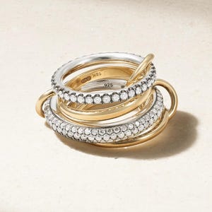 Op de afbeelding: Een set in elkaar grijpende ringen in goud- en zilverkleur. Sommige ringen zijn van glad gepolijst goud, terwijl andere zijn bezet met kleine heldere steentjes. De binnenkant van de ringen is voorzien van de gravures 18K en .925.