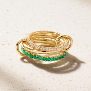 May include: A gold ring set consisting of three interlocking bands. One band features a row of clear pavé stones, another band is plain polished gold, and the third band is set with a continuous row of vibrant green circular stones.