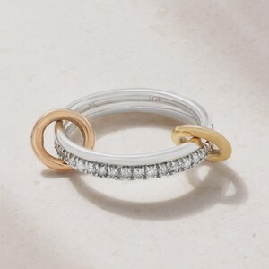 May include: A silver ring featuring a band embedded with small clear stones. The ring is accented by two attached circular links, one in a rose gold tone and one in a yellow gold tone. The interior of the silver band is stamped with the text 925.