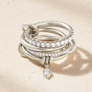 Op de afbeelding: Een zilverkleurige ring met meerdere banden bestaande uit diverse in elkaar grijpende, gladde metalen ringen. Twee van de banden zijn bezet met kleine, heldere, rondgeslepen stenen. Een enkele marquise-geslepen steen hangt in het midden.