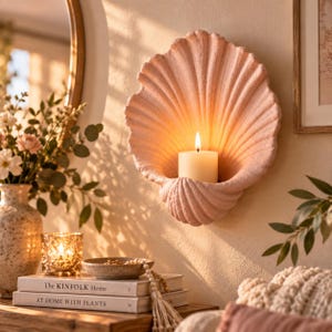 Puede incluir: Un portavelas de pared con forma de concha marina en color rosa pálido y superficie texturizada. Una vela de pilar blanca se sitúa en el centro de la concha, emitiendo una luz cálida y brillante que ilumina las estrías de la estructura.