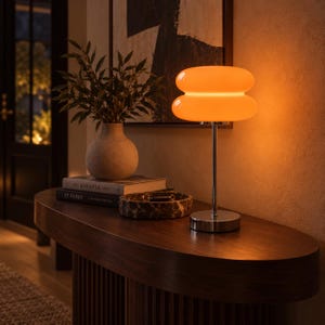 May include: A table lamp featuring a double-tiered, glowing orange glass shade supported by a slender, polished chrome metal stem and a circular base. The lamp emits a warm, diffused light, illuminating the surrounding area with a soft, golden hue.