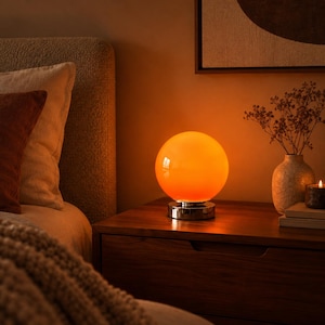Puede incluir: Una lámpara de mesa esférica de cristal naranja con una base de cromo pulido. La lámpara emite una luz cálida y brillante, proyectando un suave tono naranja sobre el área circundante. El diseño presenta una pantalla de cristal lisa y redonda.