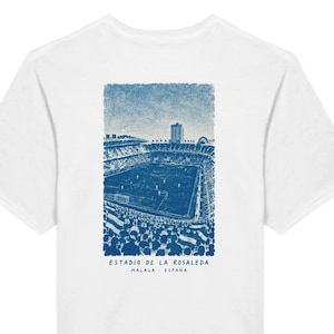 May include: A white short-sleeved t-shirt featuring a blue graphic print on the back. The illustration depicts a crowded soccer stadium with the text "ESTADIO DE LA ROSALEDA MALAGA - ESPAÑA" printed in blue lettering beneath the stadium image.