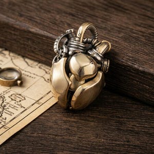May include: A metallic pendant shaped like an anatomical heart with a polished gold-tone finish and dark silver-tone mechanical details including pipes and valves. The pendant features a complex, layered design with smooth, rounded surfaces.
