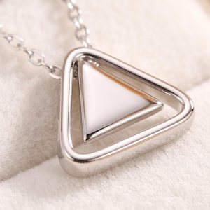 May include: A silver-toned necklace featuring a pendant with a polished triangular outer frame and a smooth, solid white triangular center inlay. The pendant is suspended from a delicate silver-toned chain link necklace.