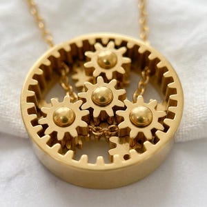 May include: A gold-toned pendant necklace featuring a circular outer gear frame containing three smaller, interconnected gear-shaped charms. The metallic design includes a central gear and two flanking gears, all suspended by a delicate gold chain.