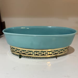 May include: An oval-shaped turquoise bowl with a smooth finish, set inside a decorative gold-toned metal stand. The stand features a repeating geometric rectangular pattern and is supported by small, short legs at the base of the structure.