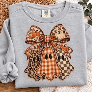 Op de afbeelding: Een lichtgrijs sweatshirt met ronde hals en een grote afbeelding van een decoratieve strik met pompoenen en herfstbladeren boven drie geesten. De geesten hebben verschillende patronen: luipaardprint, oranje gingham en een bruin-wit schaakbordpatroon.