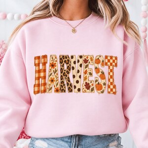 May include: A light pink crewneck sweatshirt featuring the word HARVEST printed across the chest. Each letter displays a unique pattern including orange plaid, floral prints, leopard spots, brown polka dots, autumn leaves, and pumpkins.