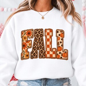 Peut inclure: Un sweat-shirt blanc à col rond avec le mot FALL écrit en grandes lettres majuscules. Chaque lettre présente un motif distinct : le F a des tournesols orange, le A un imprimé léopard, le L un damier orange et le L des citrouilles.