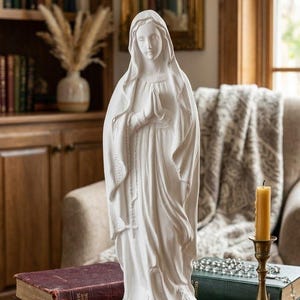 Könnte beinhalten: Eine weiße, matte Statue der Jungfrau Maria, die mit gefalteten Händen betet und ein drapiertes Gewand sowie einen Schleier trägt. Die Figur steht auf einem runden Holzsockel und zeigt detaillierte Stofffalten sowie einen seitlichen Rosenkranz.