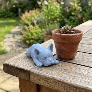 Può includere: Una statuetta di bulldog francese color azzurro in posizione sdraiata con la testa appoggiata sulle zampe anteriori, posizionata accanto a un piccolo vaso di terracotta contenente una pianta succulenta su una superficie in legno.