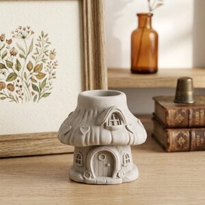 May include: A light gray, mushroom-shaped house figurine with a textured roof, heart-shaped indentations, a central arched door, and small windows. The structure features a hollow top opening and a detailed, carved exterior surface design.